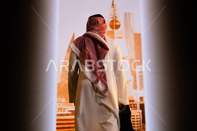 تنفيذ مشاريع تنموية عمرانية في السعودية, رؤية سعودية 2030, الأبراج و الإنجازات العمرانية في الرياض , الافتخار بالإنجازات التي تحققت بفضل توحيد البلاد, صورة من الخلف لرجل عربي سعودي خليجي يرتدي الشماغ والثوب التقليدي و يمسك حقيبة يد ويقف امام أبراج الرياض