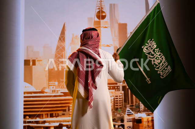 استلهام روح الإرادة والعزيمة، اليوم الوطني لتوحيد المملكة 8 ربيع الاول، العيد الوطني السعودي, تضامن ووحدة السعوديين باحتفالاتهم بيوم العلم 11 مارس, صورة من الخلف لرجل عربي سعودي خليجي يرتدي الشماغ والثوب التقليدي ويحمل علم المملكة ويقف امام أبراج الرياض