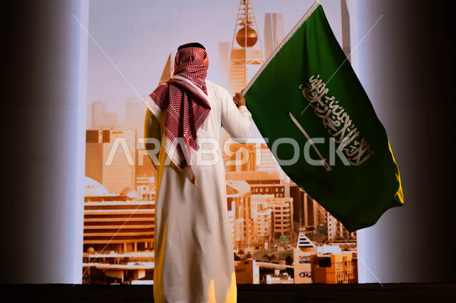 تضامن ووحدة السعوديين باحتفالاتهم بيوم العلم 11 مارس, العيد الوطني السعودي, استلهام روح الإرادة والعزيمة باستذكار اليوم الوطني 8 ربيع الاول، صورة من الخلف لرجل عربي سعودي خليجي يرتدي الشماغ والثوب التقليدي ويحمل علم المملكة ويقف امام أبراج الرياض