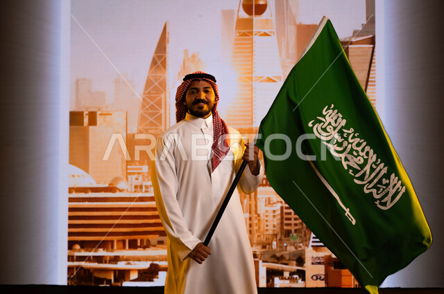 العيد الوطني السعودي 23 سبتمبر, تضامن ووحدة السعوديين باحتفالاتهم بيوم العلم 11 مارس, استلهام روح الإرادة والعزيمة باستذكار اليوم الوطني لتوحيد المملكة، رجل عربي سعودي خليجي يرتدي الزي التقليدي ويحمل علم المملكة, خلفية أبراج وناطحات سحاب مدينة الرياض
