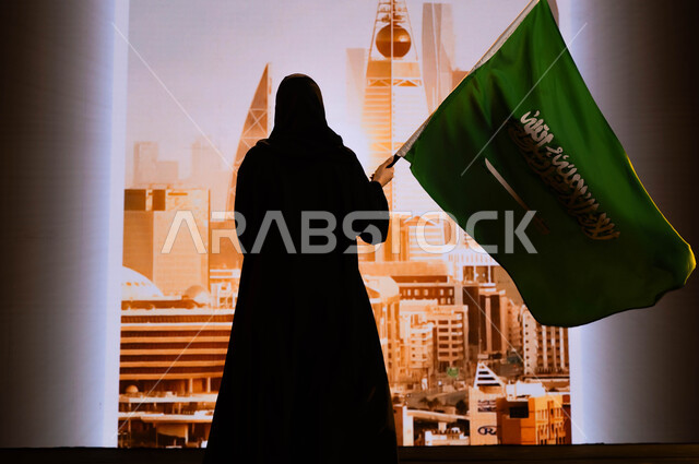 Commemorating the history of our ancestors and celebrating their achievements, the National Day, September 23, the celebration of the Saudi Flag Day, March 11, the anniversary of the Saudi National Day, a picture from behind of a Saudi Gulf Arab woman wearing the black abaya, carrying the flag of the Kingdom, and standing in front of the towers and skyscrapers of the city of Riyadh.