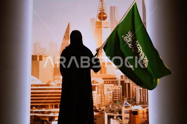Commemoration of National Day September 23, Saudi National Day, celebration of Saudi Flag Day March 11, a picture from behind of a Saudi Gulf Arab woman wearing the black abaya, carrying the flag of the Kingdom and standing in front of the towers and skyscrapers of the city of Riyadh, commemorating the history of the ancestors and celebrating their achievements