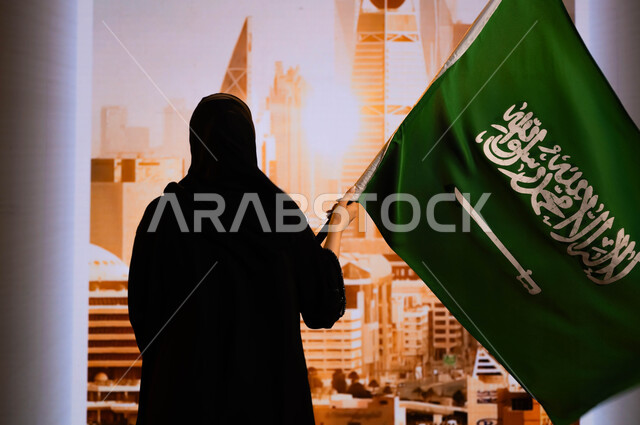 الاحتفال بيوم العلم السعودي 11 مارس, العيد الوطني السعودي, ذكرى اليوم الوطني 23 سبتمبر, صورة من الخلف لإمرأة عربية سعودية خليجية ترتدي العباءة السوداء وتحمل علم المملكة وتقف أمام ابراج وناطحات سحاب مدينة الرياض, إحياء تاريخ الأجداد والاحتفال بإنجازاتهم
