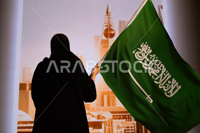 هوية اليوم الوطني السعودي, الاحتفال بيوم العلم 11 مارس, ذكرى اليوم الوطني 23سبتمبر, صورة من الخلف لإمرأة عربية سعودية خليجية ترتدي العباءة السوداء وتحمل علم المملكة وتقف امام ابراج وناطحات سحاب الرياض, إحياء تاريخ الأجداد والاحتفال بإنجازاتهم