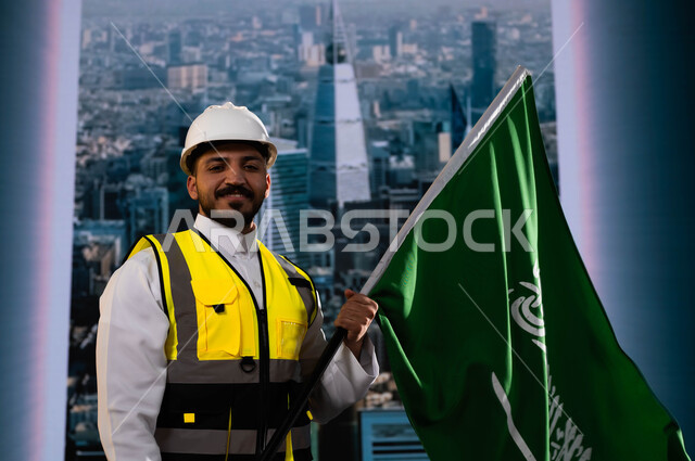 نهضة الوطن بفضل أبنائه, العيد الوطني السعودي 8 ربيع الاول, اليوم الوطني لذكرى توحيد السعودية, الاحتفال بيوم العلم 11 مارس, صورة مقربة لمهندس عربي سعودي خليجي يرتدي الخوذة وسترة الحماية ويمسك علم المملكة, خلفية أبراج الرياض المتميزة