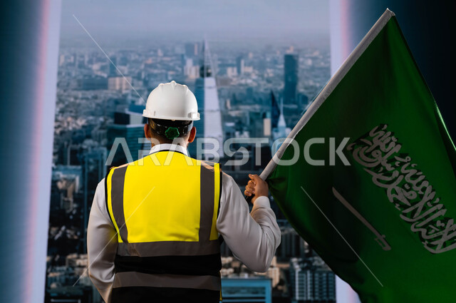 Commemorating the date of the National Day, September 23, the National Day commemorating the unification of Saudi Arabia, celebrating Flag Day, March 11, a picture from behind of a Saudi-Gulf Arab engineer wearing a helmet and a protective vest, holding the Kingdom’s flag and standing in front of the towers and skyscrapers of Riyadh, the nation’s renaissance thanks to its sons