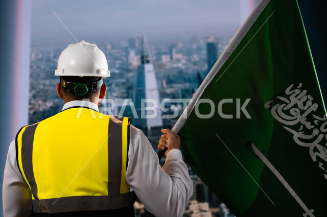 تخليد تاريخ توحيد السعودية 23 سبتمبر، اليوم الوطني السعودي, الاحتفال بيوم العلم 11 مارس، صورة من الخلف لمهندس عربي سعودي خليجي يرتدي الخوذة وسترة الحماية ويمسك علم المملكة، الوقوف امام أبراج الرياض المتميزة، نهضة الوطن بفضل أبنائه