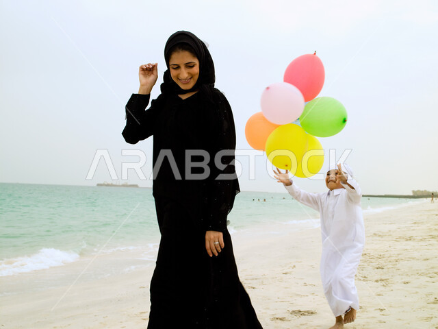 The importance of sharing leisure time with children, running and jogging on the seashore, happy family memories and moments, an Arab Emirati mother wearing an abaya playing with balloons with her son on the soft sands of the sea, tourism places and areas