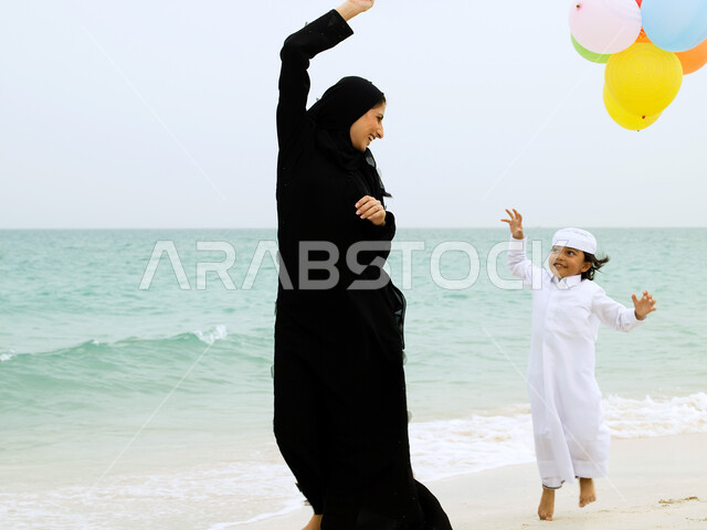 أهمية مشاركة الوقت الترفيهي مع الأبناء, الجري والركض على شاطئ البحر، الذكريات واللحظات العائلية السعيدة, أم عربية خليجية إماراتية ترتدي العباءة تلعب بالبلالين مع ابنها على رمال البحر الناعمة, أماكن ومناطق السياحة في الخليج, منتجعات شاطئية إماراتية.