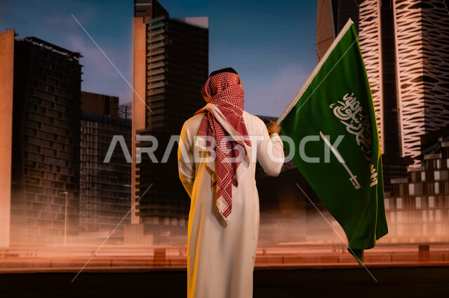 يوم العلم السعودي 11مارس, تجديد ذكرى توحيد المملكة 8 ربيع الاول, مفهوم الولاء للوطن والاحتفال باليوم الوطني, تنمية روح الانتماء للوطن, صورة من الخلف لرجل عربي سعودي خليجي يرتدي الشماغ والثوب التقليدي ويحمل علم المملكة ويقف امام مركز الملك عبد الله المالي