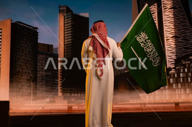 تنمية روح الانتماء للوطن، تجديد ذكرى توحيد المملكة 8 ربيع الاول، مفهوم الولاء للبلاد والاحتفال باليوم الوطني السعودي، يوم العلم السعودي 11مارس، صورة من الخلف لرجل عربي سعودي خليجي يرتدي الزي التقليدي ويحمل علم المملكة ويقف امام مركز الملك عبد الله المالي