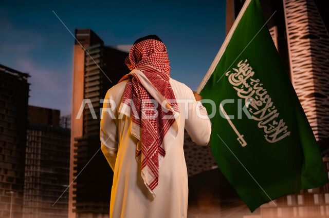 تجديد ذكرى اليوم الوطني لتوحيد المملكة 8 ربيع الاول، مفهوم الولاء للبلاد والاحتفال بالعيد الوطني، يوم العلم السعودي 11مارس، صورة من الخلف لرجل عربي سعودي خليجي يرتدي الشماغ والثوب التقليدي ويحمل علم السعودية ويقف امام ابراج مركز الملك عبد الله المالي