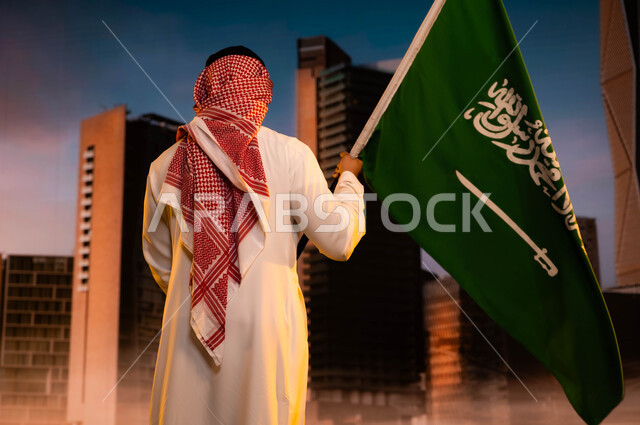 تجديد ذكرى اليوم الوطني لتوحيد السعودية 8 ربيع الاول، مفهوم الولاء والانتماء للوطن والاحتفال بالعيد الوطني، يوم العلم السعودي 11مارس، صورة من الخلف لرجل عربي سعودي خليجي يرتدي الشماغ والثوب التقليدي ويحمل علم المملكة ويقف امام مركز الملك عبد الله المالي