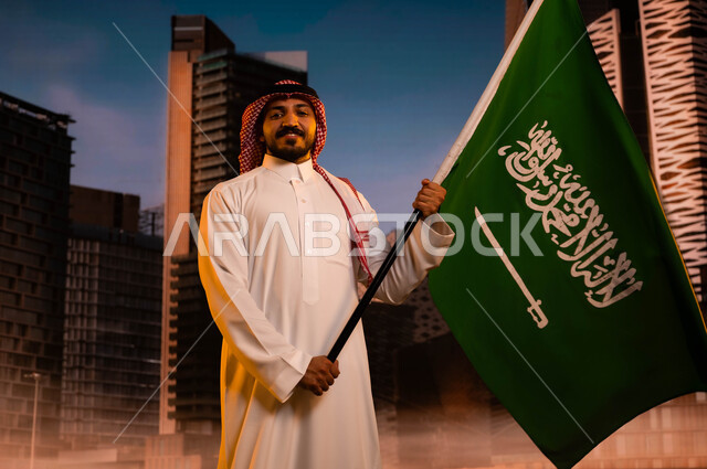 الاحتفال باليوم الوطني السعودي، تجديد الذكرى التاريخية لتوحيد المملكة، يوم العلم السعودي 11مارس، تنمية روح الانتماء للوطن، رجل عربي سعودي خليجي يرتدي الشماغ والثوب التقليدي ويحمل علم السعودية، العيد الوطني 23 سبتمبر، خلفية مركز الملك عبد الله المالي