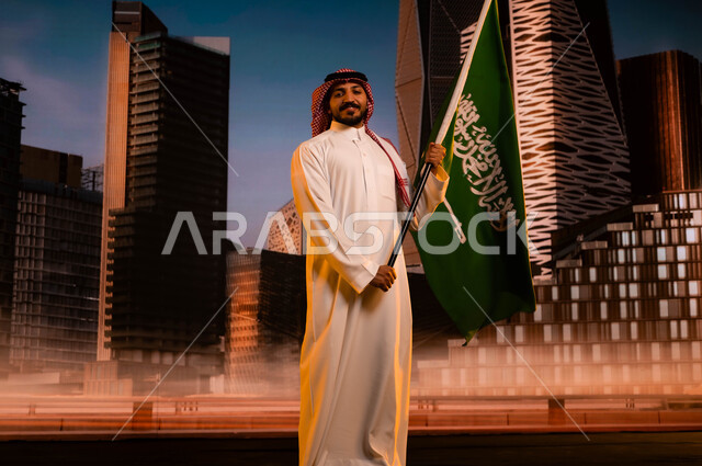 الذكرى التاريخية لتوحيد المملكة 23 سبتمبر، الاحتفال باليوم الوطني السعودي، يوم العلم السعودي 11مارس، تنمية روح الانتماء للوطن, رجل عربي سعودي خليجي يرتدي الشماغ والثوب التقليدي ويحمل علم السعودية، العيد الوطني السعودي، خلفية مركز الملك عبد الله المالي