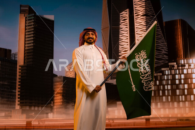 الذكرى التاريخية لتوحيد المملكة 23 سبتمبر, مفهوم الانتماء للوطن والاحتفال باليوم الوطني السعودي، يوم العلم السعودي 11مارس، تنمية روح الانتماء للوطن، رجل عربي سعودي خليجي يرتدي الشماغ والثوب التقليدي ويحمل علم السعودية، خلفية مركز الملك عبد الله المالي