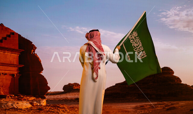 مفهوم الوطنية بالاحتفال بيوم العلم 11 مارس، استرجاع ذكرى اليوم الوطني السعودي، استلهام روح الإرادة والعزيمة باستذكار اليوم الوطني للمملكة 8 ربيع الاول، صورة من الخلف لرجل عربي سعودي خليجي يرتدي الزي التقليدي ويحمل علم المملكة ويقف أمام قصر الفريد في العلا