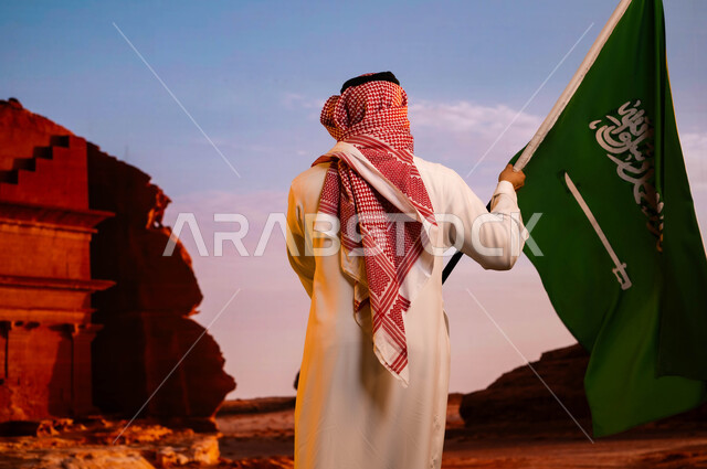 استلهام روح الإرادة والعزيمة واستذكار العيد الوطني 8 ربيع الاول، وحدة السعوديين وتضامنهم باحتفالهم بيوم العلم 11 مارس، ذكرى اليوم الوطني السعودي، صورة من الخلف لرجل عربي سعودي خليجي يرتدي الزي التقليدي ويحمل علم المملكة ويقف أمام قصر الفريد في العلا