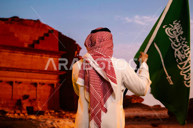 ذكرى العيد الوطني السعودي، وحدة السعوديين وتضامنهم باحتفالهم بيوم العلم 11 مارس، استلهام روح الإرادة والعزيمة، اليوم الوطني السعودي 8 ربيع الاول، صورة من الخلف لرجل عربي سعودي خليجي يرتدي الزي التقليدي ويحمل علم المملكة ويقف امام قصر الفريد في العلا