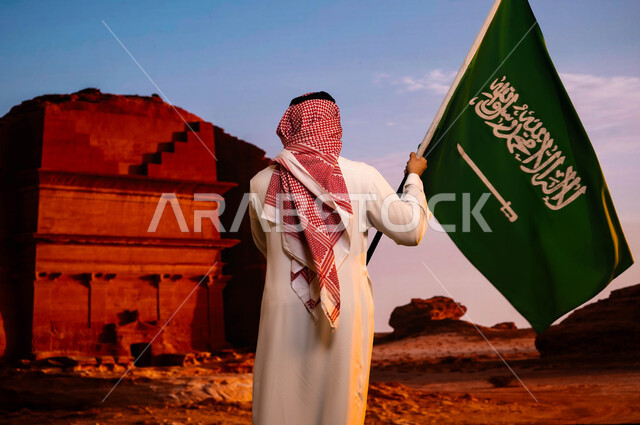 وحدة السعوديين وتضامنهم باحتفالهم بيوم العلم 11 مارس، ذكرى اليوم الوطني السعودي، استلهام روح الإرادة والعزيمة، اليوم الوطني لتوحيد المملكة 8 ربيع الاول، صورة من الخلف لرجل عربي سعودي خليجي يرتدي الزي التقليدي ويحمل علم المملكة وينظر لقصر الفريد في العلا