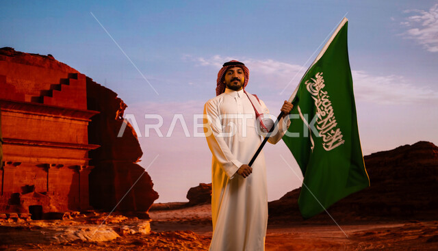 إحياء ذكرى يوم العلم 11 مارس، تضامن ووحدة السعوديين باحتفالاتهم باليوم الوطني السعودي، استلهام روح الإرادة والعزيمة باستذكار يوم توحيد المملكة 23 سبتمبر، رجل عربي سعودي خليجي يرتدي الشماغ والثوب التقليدي ويحمل علم المملكة، خلفية قصر الفريد في منطقة العلا