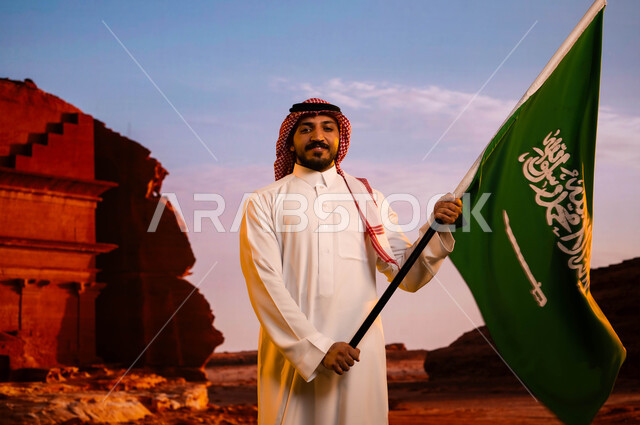 تضامن ووحدة السعوديين باحتفالاتهم بيوم العلم 11 مارس، ذكرى اليوم الوطني السعودي، استلهام روح الإرادة والعزيمة، العيد الوطني لتوحيد المملكة 23 سبتمبر، رجل عربي سعودي خليجي يرتدي الشماغ والثوب التقليدي ويحمل علم المملكة، خلفية قصر الفريد في منطقة العلا