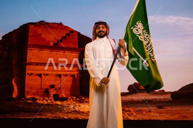العيد الوطني السعودي، تضامن ووحدة السعوديين باحتفالاتهم بيوم العلم 11 مارس، استلهام روح الإرادة والعزيمة باستذكار اليوم الوطني لتوحيد المملكة 23 سبتمبر، رجل عربي سعودي خليجي يرتدي الشماغ والثوب التقليدي ويحمل علم المملكة، خلفية قصر الفريد في منطقة العلا