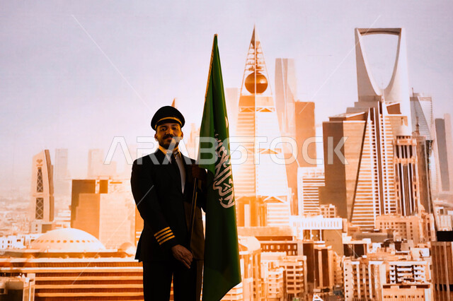 ذكرى اليوم الوطني لتوحيد المملكة على يد الملك عبد العزيز، إيماءات الاعتزاز بإنجازات الوطن، طيار عربي سعودي خليجي يرتدي بدلة الطيران ويقف بشموخ ويحمل علم السعودية، الاحتفال بالعيد الوطني السعودي 23 سبتمبر، يوم العلم 11 مارس، خلفية أبراج مدينة الرياض