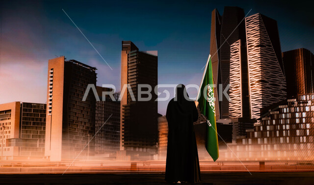 مفهوم الولاء والانتماء للوطن، العيد الوطني لذكرى توحيد المملكة في 23سبتمبر، الاحتفال باليوم الوطني السعودي، صورة من الخلف لامرأة عربية خليجية سعودية محجبة تحمل علم السعودية وتقف امام أبراج مركز الملك عبد الله المالي في الرياض، يوم العلم السعودي 11 مارس