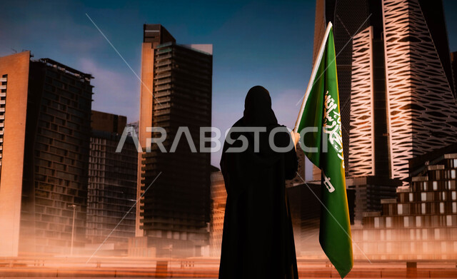 مفهوم حب الوطن، العيد الوطني لتوحيد المملكة في 8 ربيع الاول، الاحتفال باليوم الوطني السعودي، صورة من الخلف لامرأة عربية خليجية سعودية محجبة ترتدي العباءة وترفع علم السعودية وتقف أمام أبراج مركز الملك عبد الله المالي (كافد) في الرياض، يوم العلم 11 مارس