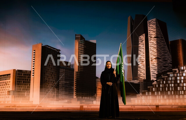 الاحتفال باليوم الوطني السعودي 23 سبتمبر، ترسيخ جذور الانتماء للوطن، امرأة عربية خليجية سعودية محجبة ترتدي العباءة وتحمل علم السعودية، يوم العلم السعودي 11 مارس، ذكرى توحيد المملكة على يد الملك عبد العزيز، خلفية أبراج مركز الملك عبد الله المالي (كافد)