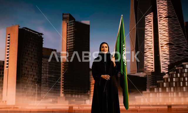 ترسيخ جذور الانتماء للوطن، الاحتفال باليوم الوطني السعودي، امرأة عربية خليجية سعودية محجبة ترتدي العباءة وتحمل علم السعودية، يوم العلم السعودي 11 مارس، ذكرى توحيد المملكة على يد الملك عبد العزيز 8 ربيع الأول، خلفية أبراج مركز الملك عبد الله المالي (كافد)