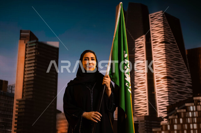 مفهوم الانتماء للوطن، الاحتفال باليوم الوطني السعودي، صورة مقربة لامرأة عربية خليجية سعودية محجبة ترتدي العباءة وتحمل علم السعودية، العيد الوطني لذكرى توحيد الملك عبد العزيز للمملكة في 23 سبتمبر، خلفية أبراج مركز الملك عبد الله المالي(كافد)