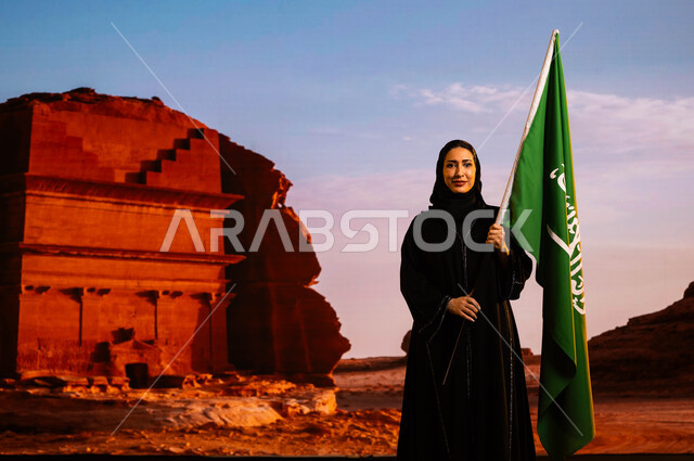 Commemoration of the Saudi National Day, Saudi national holidays and events, National Day 8 Rabi’ al-Awwal, Flag Day 11 March, a smiling Saudi Gulf Arab woman wearing the black abaya and carrying the flag of the Kingdom, commemorating the history of the ancestors and celebrating their achievements, the background of the historic Al-Farid Palace in Al-Ula