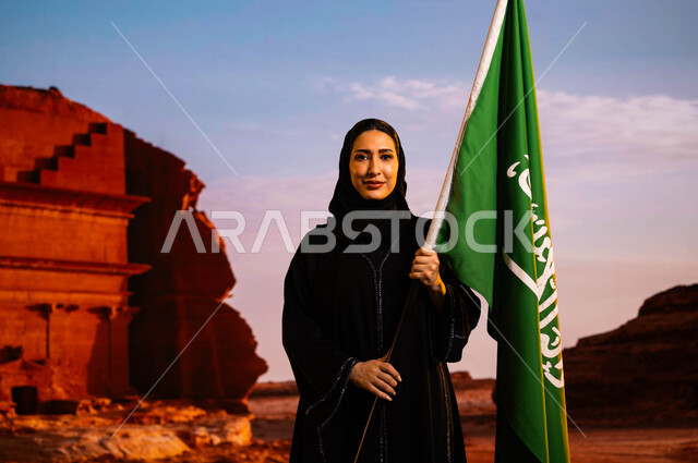 Commemorating the history of our ancestors and celebrating their achievements, Saudi national holidays and events, celebrating the anniversary of the 8th Rabi’ al-Awwal National Day, Saudi National Day, Flag Day March 11, a smiling Saudi Gulf Arab woman wearing the black abaya and carrying the flag of the Kingdom, the background of the Alfred Palace in Al-Ula