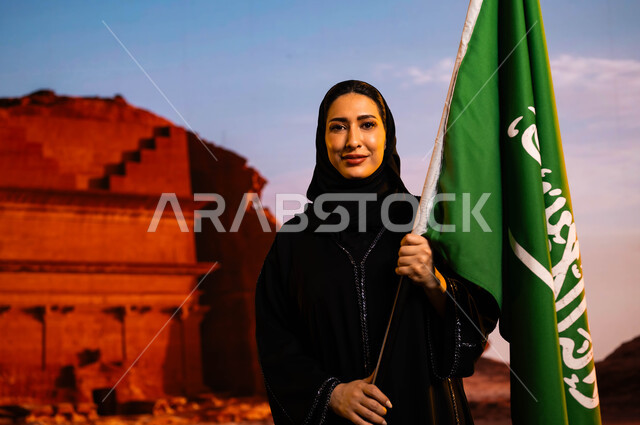 مناسبات وأعياد سعودية، الاحتفال بذكرى اليوم الوطني 8 ربيع الأول، العيد الوطني السعودي، يوم العلم 11 مارس، صورة مقربة لإمرأة عربية سعودية خليجية ترتدي العباءة السوداء وتحمل علم المملكة، إحياء تاريخ الأجداد والاحتفال بإنجازاتهم، خلفية قصر الفريد في العلا