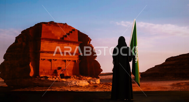 Commemorating the history of our ancestors and celebrating their achievements, Saudi National Day, celebrating Flag Day on March 11, commemorating National Day on September 23, a picture from the back of a Saudi Gulf Arab woman wearing the black abaya and standing in front of the historic Al-Farid Palace in the Al-Ula area, raising the Kingdom’s flag proudly.