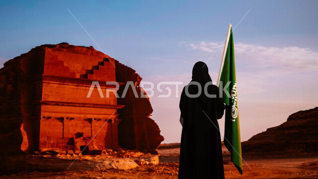 ذكرى العيد الوطني 23سبتمبر، اليوم الوطني السعودي، الاحتفال بيوم العلم 11 مارس، صورة من الخلف لإمرأة عربية سعودية خليجية ترتدي العباءة السوداء وتحمل علم المملكة، الوقوف أمام قصر الفريد في منطقة العلا، إحياء تاريخ الأجداد والاحتفال بإنجازاتهم
