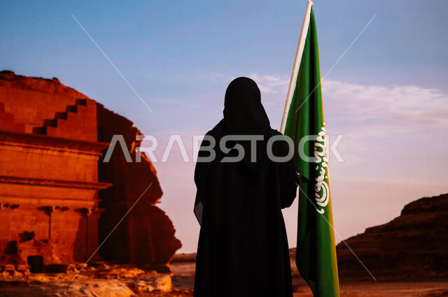 Celebrating Flag Day, March 11, Saudi National Day, commemorating National Day, September 23, a picture from the back of a Saudi Gulf Arab woman wearing the black abaya, carrying the flag of the Kingdom, standing in front of the historic Al-Farid Palace in the Al-Ula area, commemorating the history of the ancestors and celebrating their achievements