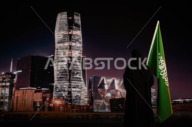 يوم العلم 11 مارس، ذكرى اليوم والعيد الوطني السعودي، صورة من الخلف لامرأة عربية خليجية سعودية محجبة ترتدي العباءة السوداء وتحمل العلم السعودي، الوقوف أمام أبراج مركز الملك عبد الله المالي(كافد)، الاحتفال بتوحيد المملكة 23 سبتمبر