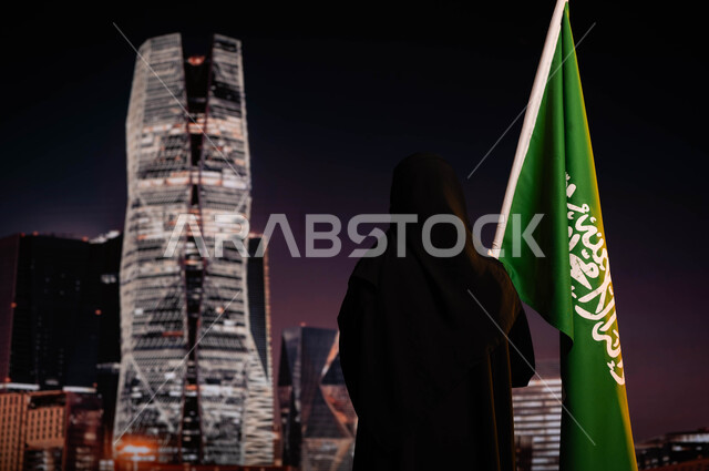 الاحتفال باليوم الوطني السعودي، يوم العلم 11 مارس، صورة من الخلف لامرأة عربية خليجية سعودية محجبة ترتدي العباءة السوداء وتقف أمام أبراج مركز الملك عبد الله المالي(كافد)، رفع علم السعودية، ذكرى توحيد المملكة 8 ربيع الأول