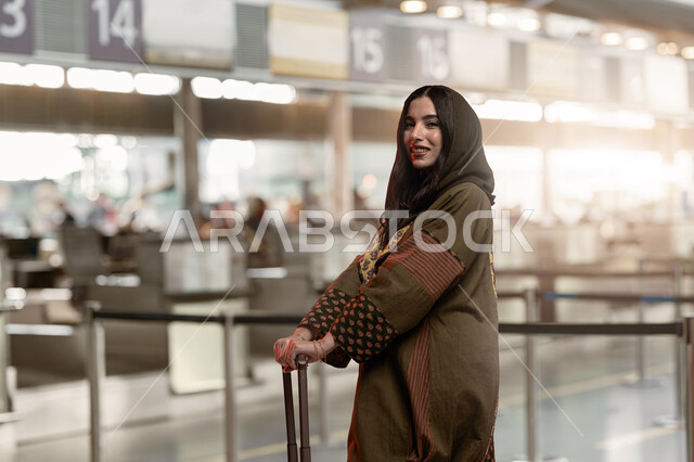 Spending fun times on the summer vacation abroad, a Saudi Arabian Gulf woman holding a suitcase in her hand, standing in the airport lounge and looking at the camera with happy gestures, preparing to depart and heading to the plane, moving around and traveling to tourist areas.