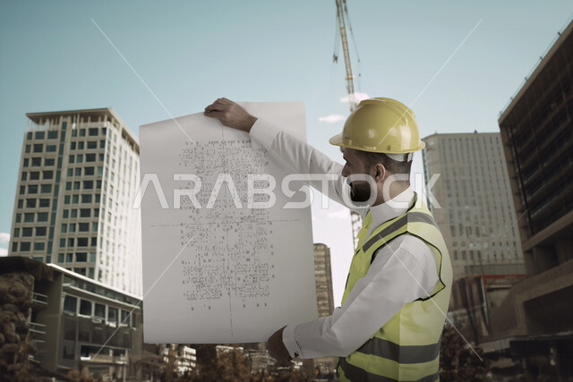 متابعة المشروع وتدقيق المخططات، مهندس معماري عربي خليجي سعودي يرتدي خوذة وسترة الوقاية الخاصة بالعمل  يحمل بيده مخطط البناء ، الاشراف والرقابة على الانشاءات الهندسية، التحقق من تطبيق المشاريع والخطط الهندسيه المعمارية في البناء بشكل صحيح في السعودية