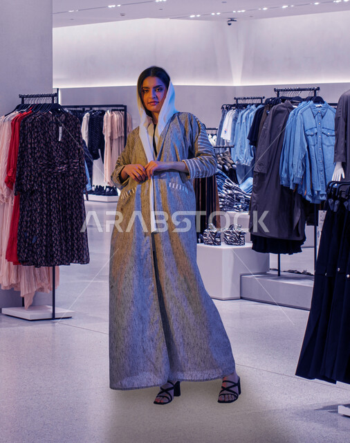 The concept of elegance and femininity, a veiled Saudi Arabian Gulf woman wearing an abaya and standing in a clothing store, taking advantage of commercial offers and discounts, enjoying buying and shopping, looking at the camera, spending an enjoyable time shopping in the mall, full-length body image
