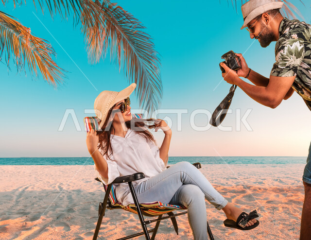 الاستمتاع بقضاء أوقات الفراغ في العطله الصيفيه أمام البحر، شاب عربي خليجي سعودي يرتدي الزي الكاجوال والنظارة الشمسية يقوم بإلتقاط صور تذكارية لشابة سعودية تجلس على الكرسي على الشاطئ في المناطق الساحليه، الترفيه عن النفس بالسياحة والسفر، أخذ وضعيات مختلفة
