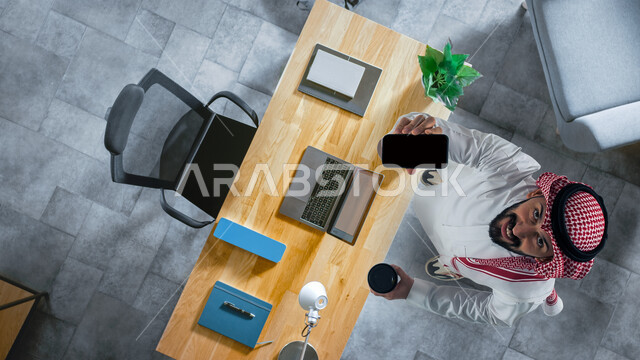 استخدام التقنيات الحديثة في الاعمال المكتبيه، صورة علوية عامودية لرجل عربي خليجي سعودي مبتسم يرتدي الثوب والزي التقليدي يقوم بعرض تطبيقات وبرامج الجوال، شاشة سوداء فارغه، الوقوف في مكتب العمل والنظر إلى الاعلى بإيماءات السعادة، تناول المشروبات الساخنة