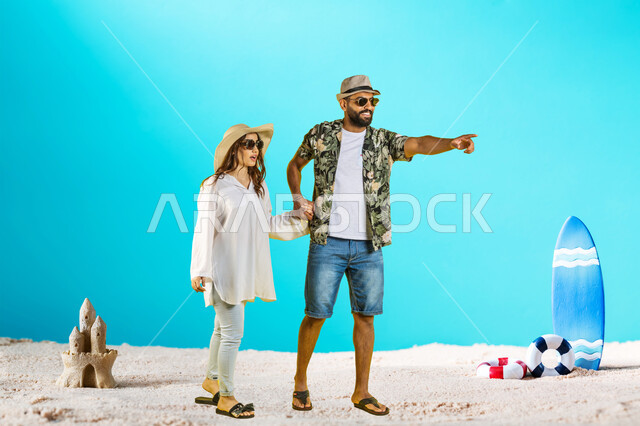 Entertaining oneself through travel and tourism, a smiling Saudi Arabian Gulf man wearing casual clothes and sunglasses with his wife standing on the soft sand and enjoying the summer vacation in the coastal areas, pointing to something, gestures of astonishment and surprise, learning about famous places.