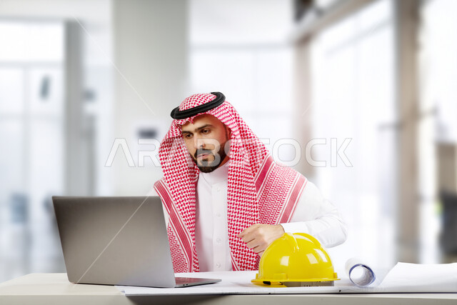 Using modern technologies to audit and evaluate architectural plans and designs, managing engineering projects using a laptop, a Saudi Gulf Arab engineer wearing a traditional dress and sitting in the work office, following up on the progress of construction projects, Saudi professions and jobs