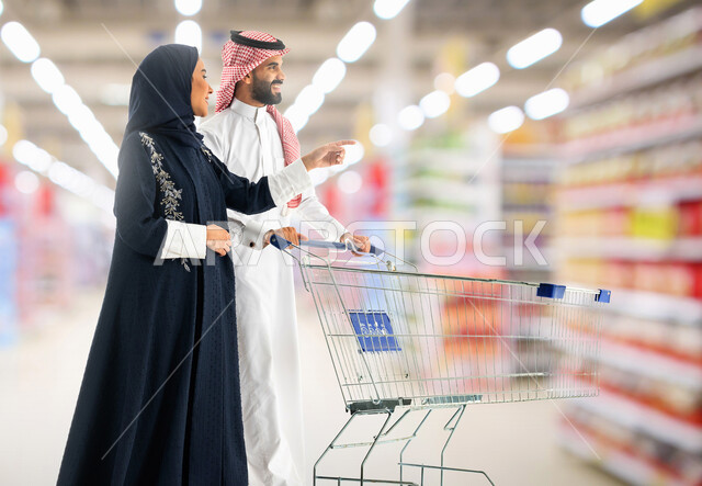 مشاركة الزوج في التبضع وشراء الإحتياجات المنزلية من السوبر ماركت، امراه عربية خليجية سعودية محجبة ترتدي العباءة الخليجيه تشير بيدها إلى شي ما، التأثير الإيجابي للتسوق على العائلة, اختيار بعض الأصناف اللازمة بإيماءات السرور، قضاء وقت ممتع في متجر المواد الغذاائية
