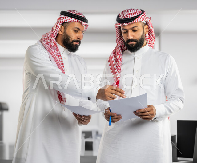 اجتماع الزملاء للمناقشة في حل المشكلات، تعزيز الروح التشاركية، توزيع المهام المطلوبة لتطوير أهداف الشركة ، رجلان توأم عربيان خليجيان سعوديان يرتديان الزي التقليدي و يحملان اوراق عمل، الوحده والعمل الجماعي، بيئة عمل سعودية